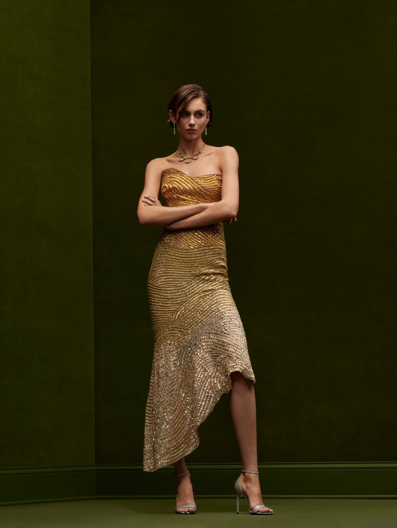 Elegant woman in a shimmering gold gown with a fishtail hem, jewelry, and metallic heels against an olive-green backdrop in a studio shot.