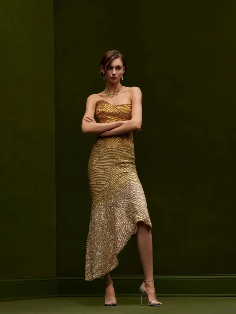 Elegant woman in a shimmering gold gown with a fishtail hem, jewelry, and metallic heels against an olive-green backdrop in a studio shot.
