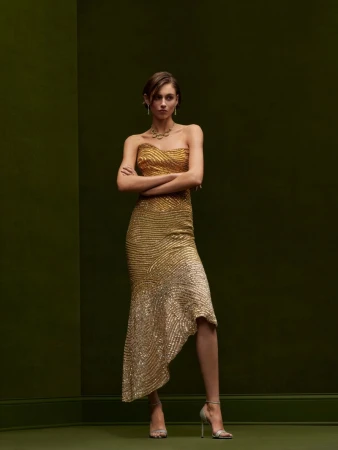 Elegant woman in a shimmering gold gown with a fishtail hem, jewelry, and metallic heels against an olive-green backdrop in a studio shot.