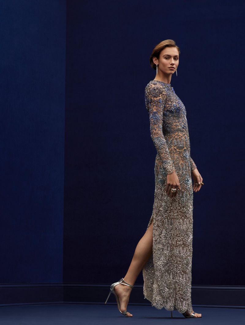 Elegant woman in a long-sleeved beaded gown and silver heels, posing against a dark blue studio backdrop with a subtle vertical color split.