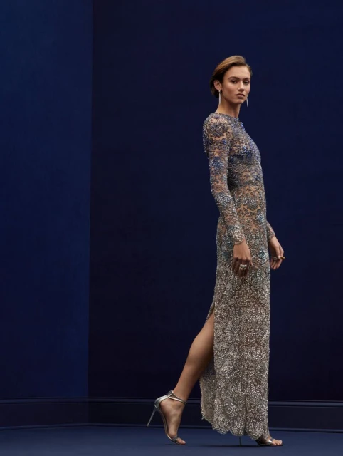 Elegant woman in a long-sleeved beaded gown and silver heels, posing against a dark blue studio backdrop with a subtle vertical color split.