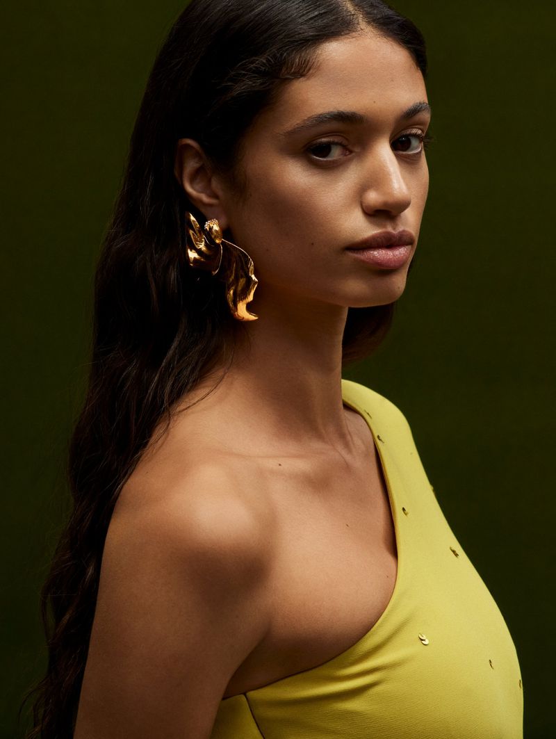 Portrait of a brunette woman with long dark hair wearing a chartreuse asymmetrical top and large gold statement earrings on a dark background.