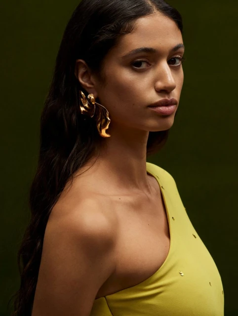Portrait of a brunette woman with long dark hair wearing a chartreuse asymmetrical top and large gold statement earrings on a dark background.