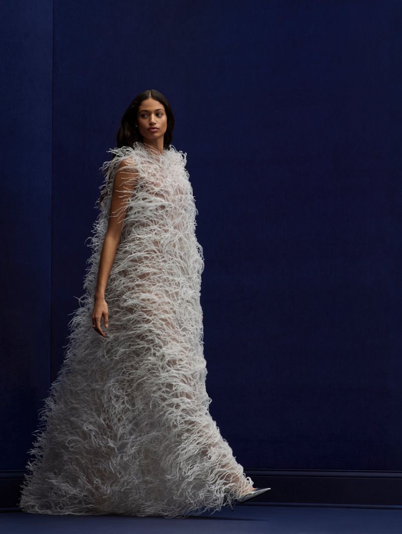 Fashion shot of an adult woman wearing a long, feathered couture dress in a photo studio with a deep blue wall and floor.