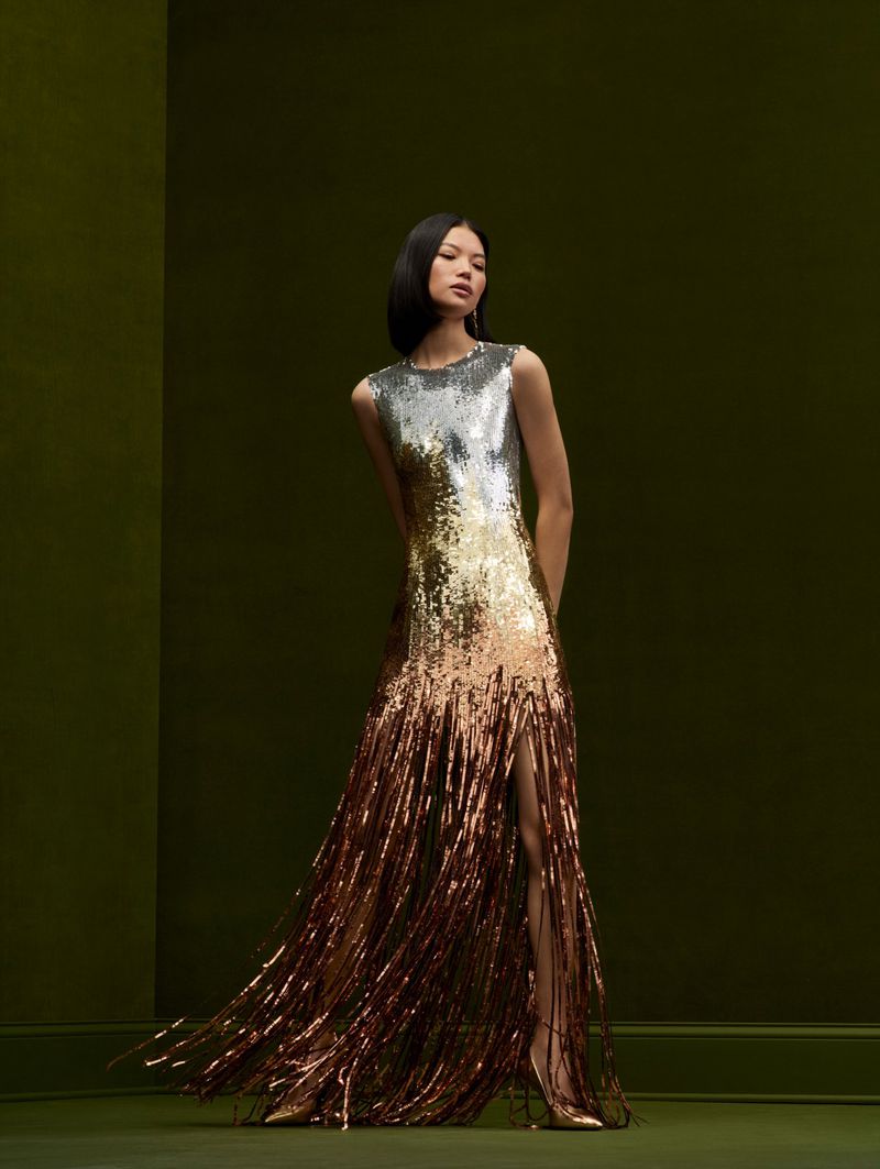 Elegant woman in a silver-to-bronze sequin gown with fringe, posing against a muted green background in a fashion forward setting.