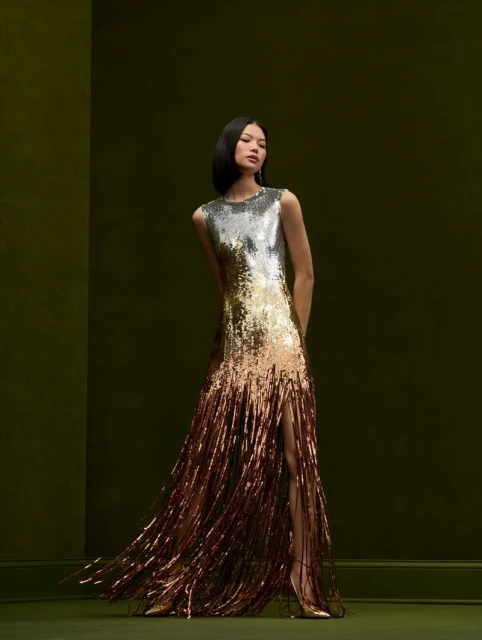 Elegant woman in a silver-to-bronze sequin gown with fringe, posing against a muted green background in a fashion forward setting.
