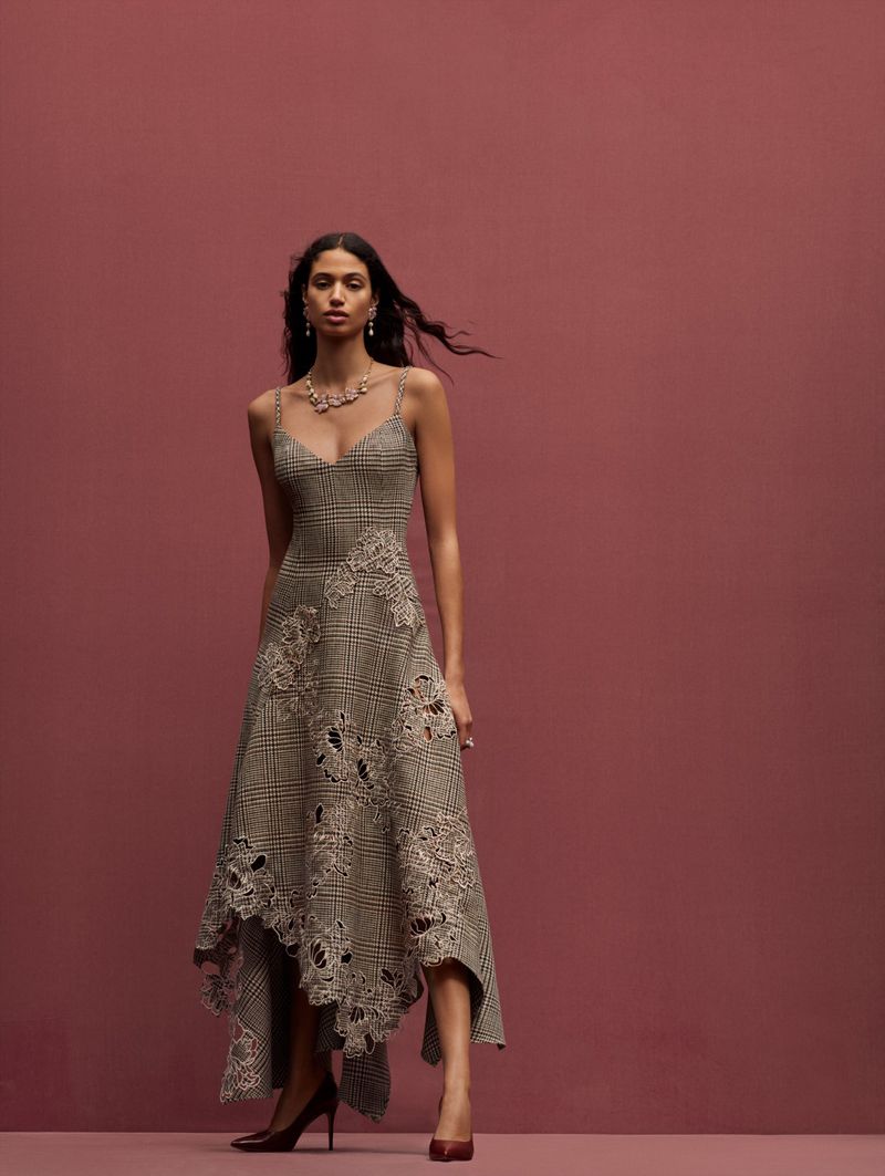 A woman stands in a sleeveless midi dress with a checked pattern, lace details and a v-neck, accessorized with jewelry, against a pink background.