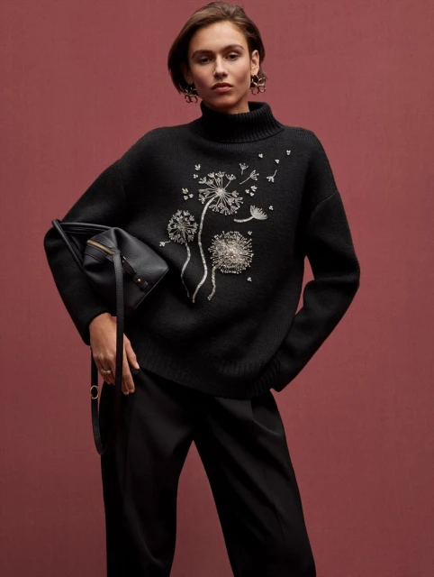 Fashion shot of a woman wearing a black turtleneck sweater with dandelion embellishments, black pants, and a black purse indoors.