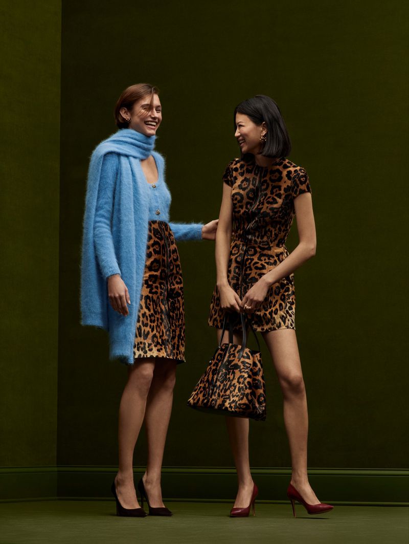Two smiling women in a photo studio pose in leopard-print dresses, one with a blue cardigan, with dark-green background and floor.