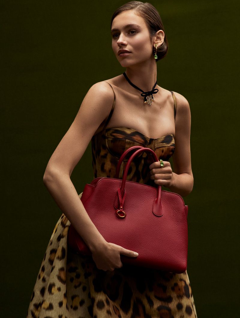 Elegant adult female in leopard print dress poses with a red leather handbag in a fashion style against a dark olive backdrop.