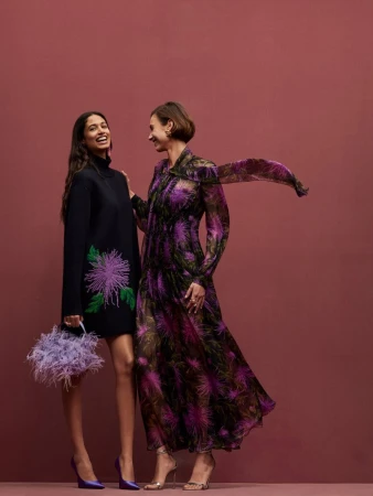 Two stylish women in floral dresses pose on a mauve background; one holds a lilac feather purse, both are smiling and looking at each other.