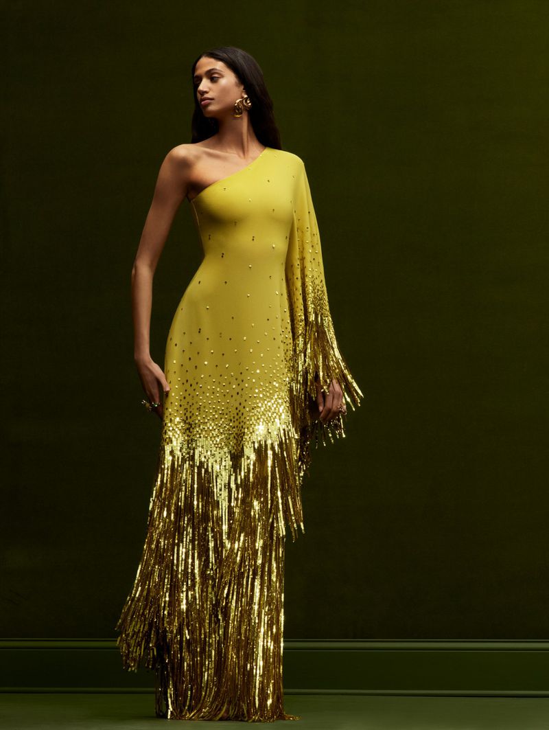 Elegant woman poses in a one-shoulder yellow gown adorned with gold fringe, set against a dark green backdrop for a fashion editorial.
