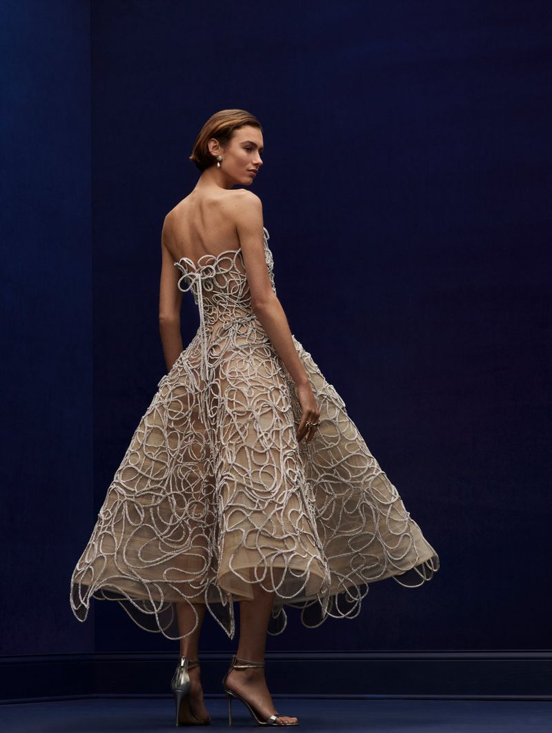 Back view of a woman in a beige strapless midi dress with white swirl details and metallic high heels against a navy blue background.