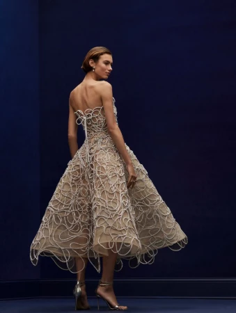 Back view of a woman in a beige strapless midi dress with white swirl details and metallic high heels against a navy blue background.