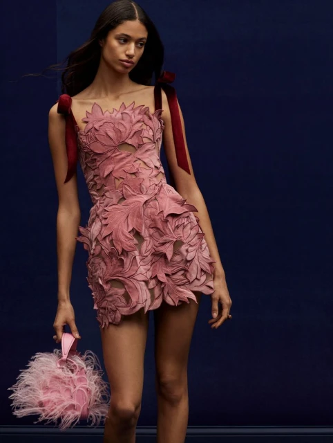 Fashion model poses in a floral-appliqued mini dress with ribbons and a feather purse against a dark blue studio backdrop.