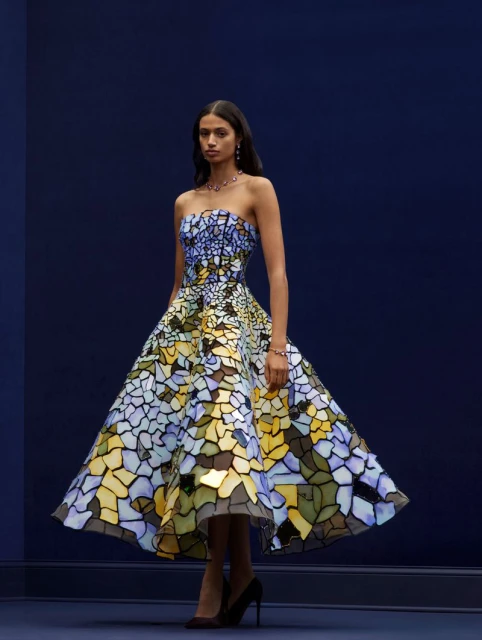 Woman in strapless floral A-line dress with dark hair poses against a dark blue studio background wearing heels and jewelry.