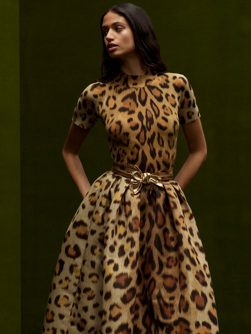 A fashionable adult female model poses in a studio wearing a leopard-print dress with a gold belt against a dark green background.