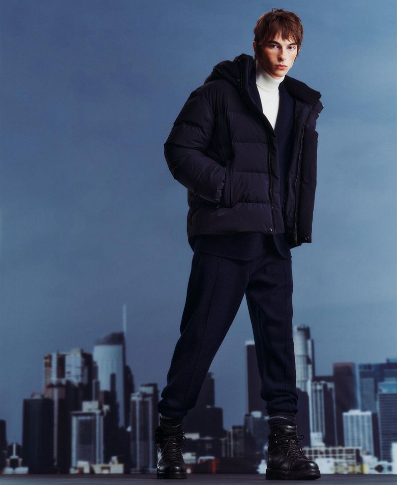 Adult male fashion model poses in a hooded puffer jacket, turtleneck, and boots against a blurred city skyline background.