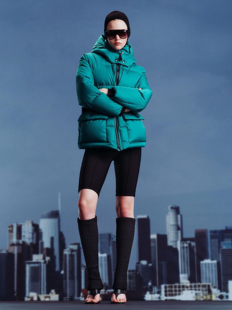 Full-length shot of a woman posing in a teal puffer coat, black cycling shorts, and knitted leg warmers against a city skyline backdrop.
