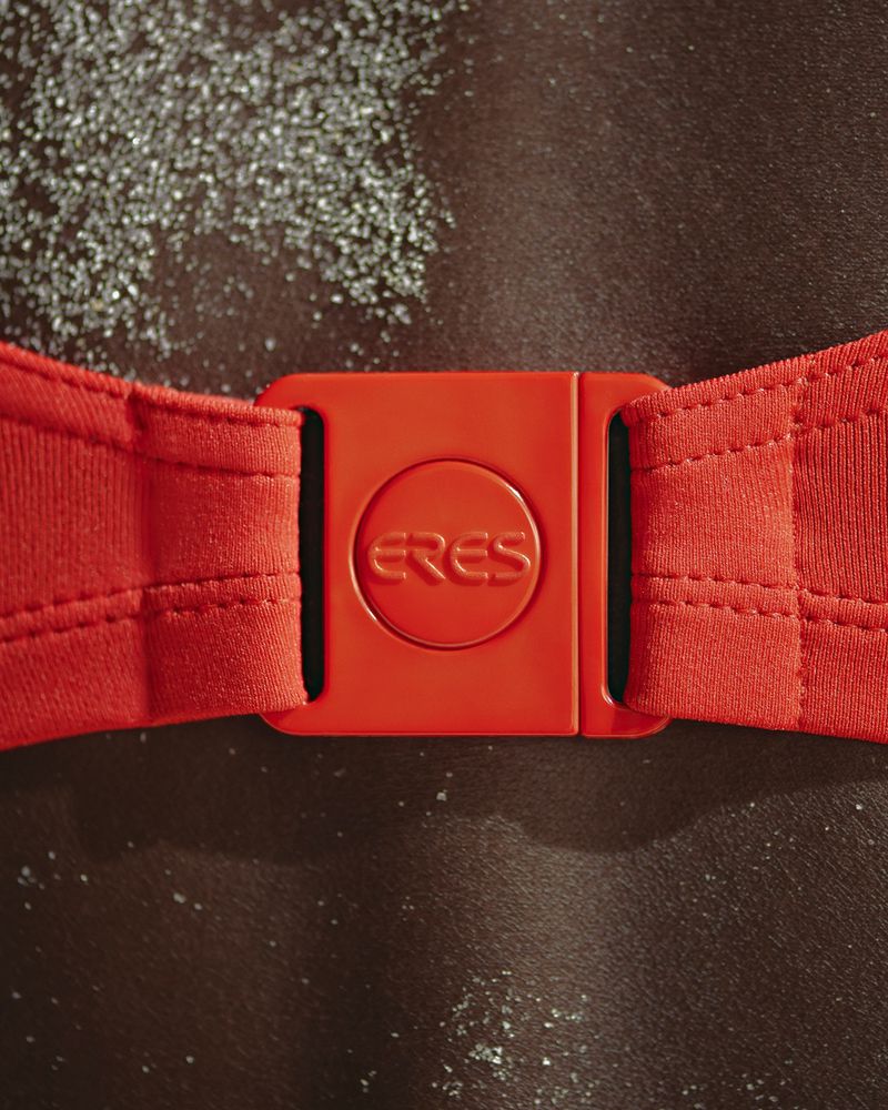 Close-up of a red swimsuit buckle with 'ERES' logo, on a dark-skinned person with sand grains. Fashion and beach-ready swimwear detail.