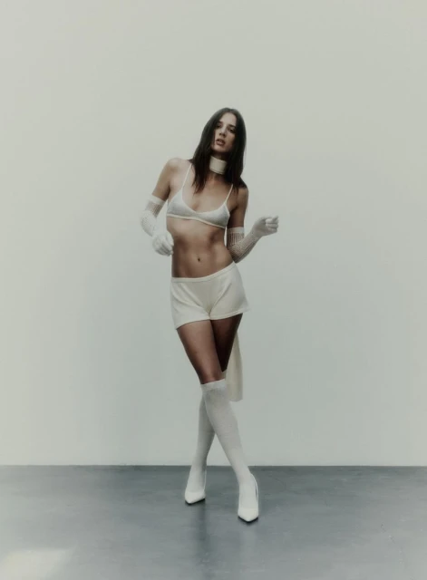 A young woman in a white bra, shorts, high socks, and heels poses in a photo studio with a blank background and studio lighting.
