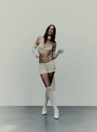 A young woman in a white bra, shorts, high socks, and heels poses in a photo studio with a blank background and studio lighting.