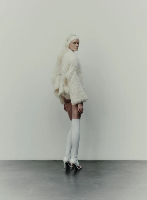 Full-length shot of a young adult woman posing in a white, textured jacket with long white socks and high heels on a gray surface.