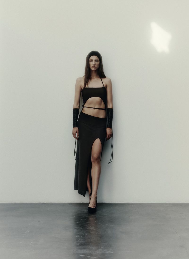 Full-length shot of a woman wearing a dark spaghetti-strap top, a high-slit skirt, and platform heels in a minimalist indoor setting.