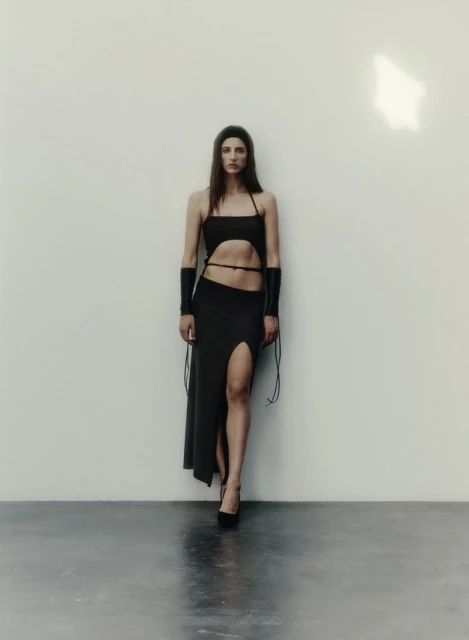 Full-length shot of a woman wearing a dark spaghetti-strap top, a high-slit skirt, and platform heels in a minimalist indoor setting.
