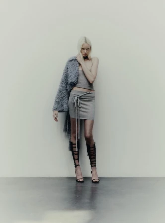 Fashion shot of an adult woman with platinum blonde hair wearing a light grey two-piece outfit and tall black gladiator sandals.