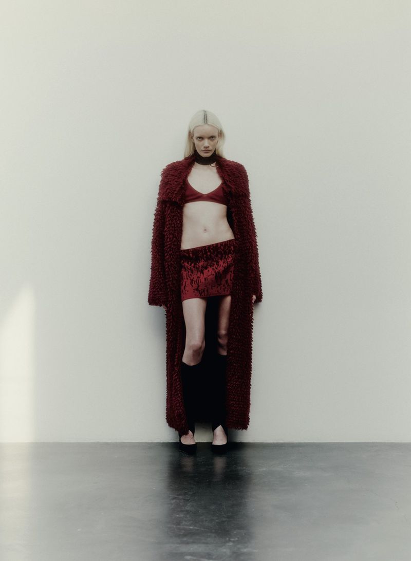 Fashion portrait of a fair-skinned adult female with light blonde hair wearing a burgundy bra top, skirt, coat and black thigh-high socks indoors.