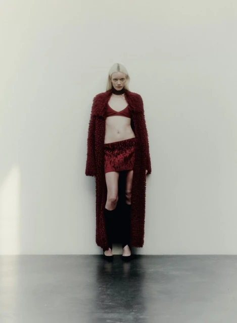 Fashion portrait of a fair-skinned adult female with light blonde hair wearing a burgundy bra top, skirt, coat and black thigh-high socks indoors.