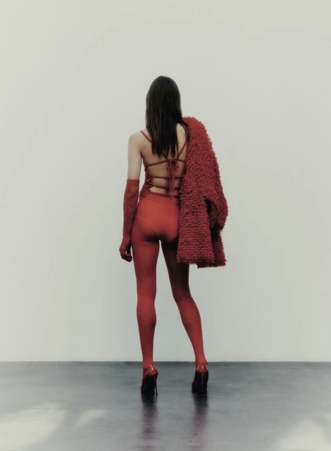 A woman with long brown hair is seen from the back, wearing a red catsuit and high heels, a red coat is hanging on her shoulder.