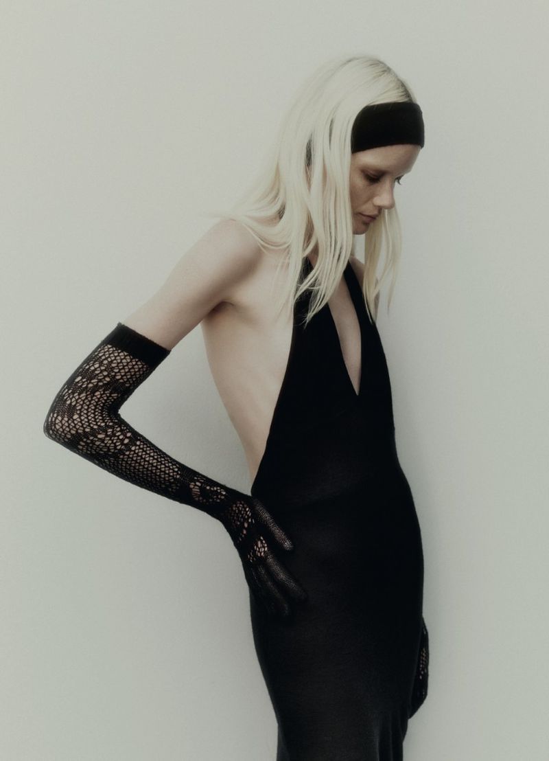 Blonde woman in low-back black halter dress and lace gloves poses against a light grey studio backdrop in a fashion shot.