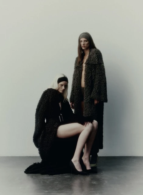 Two women wearing brown, textured coats pose in a minimalist photo studio against a solid white backdrop, highlighting fashion.