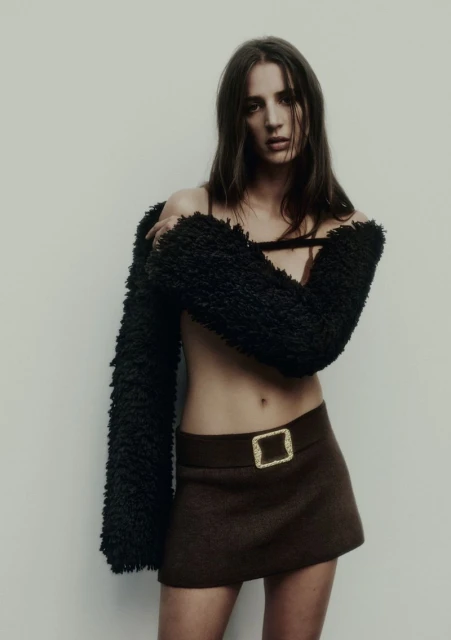 Young woman poses in brown skirt and faux fur shrug against a plain background in a fashion shot with eye-level angle.
