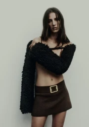 Model wearing a black fluffy textured garment and brown skirt with gold buckle belt against a neutral background