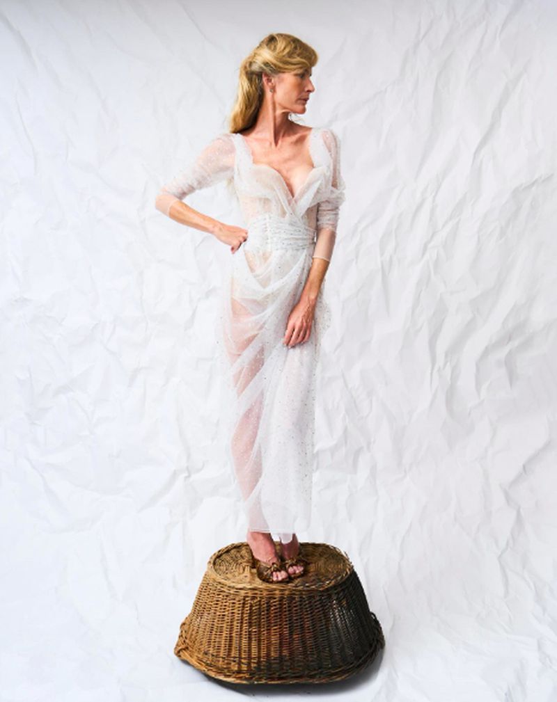 Woman in sheer white dress standing on wicker basket against a wrinkled white background in a studio fashion shot. Full length.