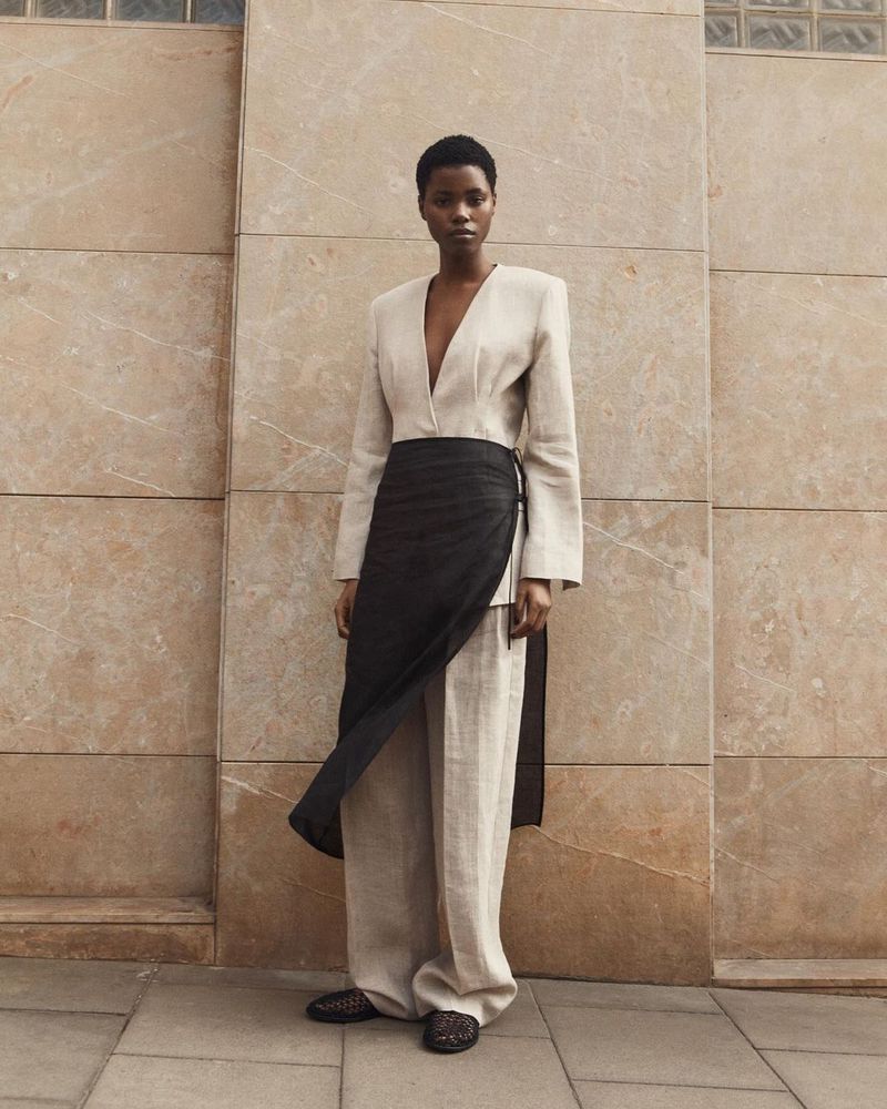 An African woman stands in neutral clothing with a black wrap, photographed in front of a textured wall and patterned shoes.