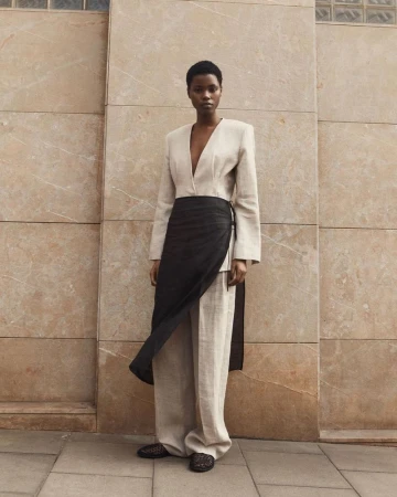 An African woman stands in neutral clothing with a black wrap, photographed in front of a textured wall and patterned shoes.