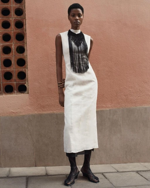 A woman in a white dress with black fringe poses against a peach wall with architectural details and wears black patterned boots.