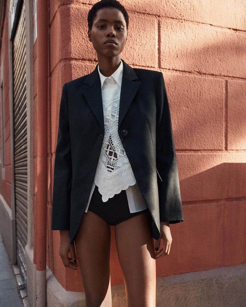 Portrait of a young Black woman wearing a black blazer and a white lace top posing in front of a textured orange wall.