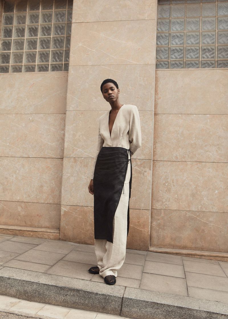 Victoria Fawole in a cream v-neck top, wide-leg trousers, and black wrap skirt standing before a stone building facade.