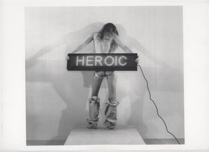 A woman stands on a platform holding a glowing sign that reads 'HEROIC', emphasizing a monochrome artistic expression.