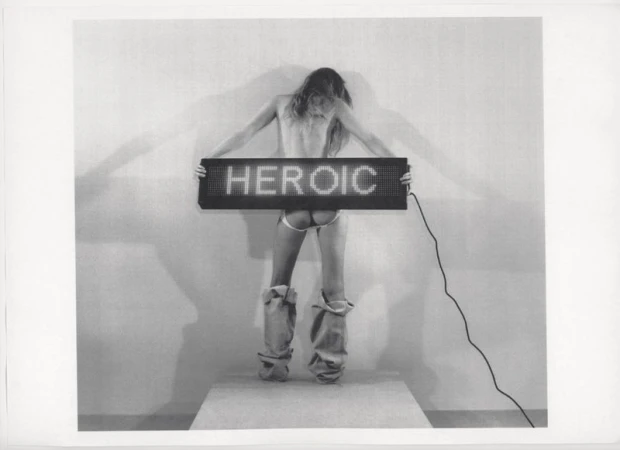 A woman stands on a platform holding a glowing sign that reads 'HEROIC', emphasizing a monochrome artistic expression.