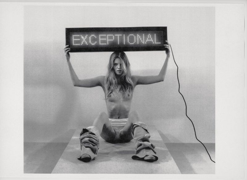 Monochrome image of a shirtless young woman holding an illuminated sign that reads 'EXCEPTIONAL' while seated indoors on a striped surface.