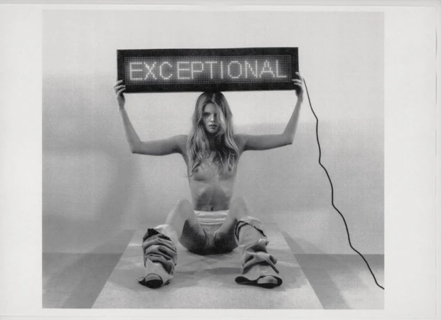 Monochrome image of a shirtless young woman holding an illuminated sign that reads 'EXCEPTIONAL' while seated indoors on a striped surface.