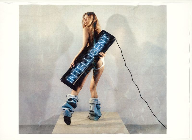 Blond woman poses with a light-up display reading 'INTELLIGENT' in a studio, wearing denim leg warmers and minimal clothing.