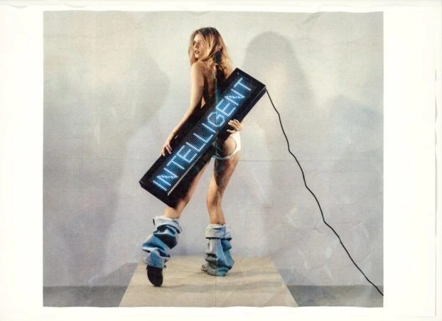 Blond woman poses with a light-up display reading 'INTELLIGENT' in a studio, wearing denim leg warmers and minimal clothing.