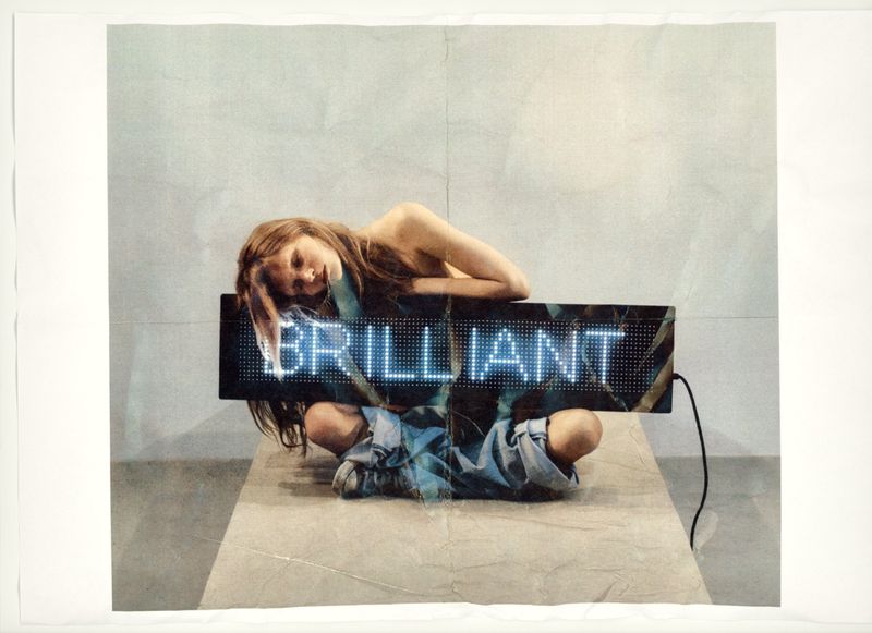 Young adult posing on a platform under a neon sign that reads 'BRILLIANT' in an indoor space with minimal background distraction.